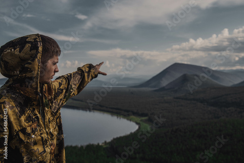 a young man in a khaki jacket stands with his back, arms outstretched, mountains