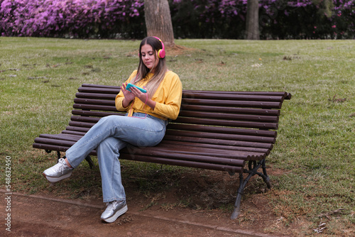 teenage girl listening to music with her headphones connected to a mobile phone on a park bench