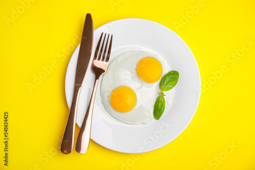 Tasty fried eggs with basil in plate on yellow background, top view