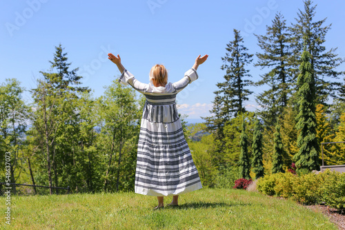 A woman in a white dress with white hair stands facing the forest with her hands up, praise and delight