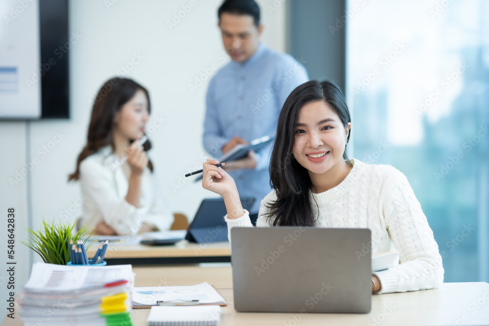 Happy beautiful Asian businesswoman sitting smiling and enjoying working on laptop in office and looking at camera.