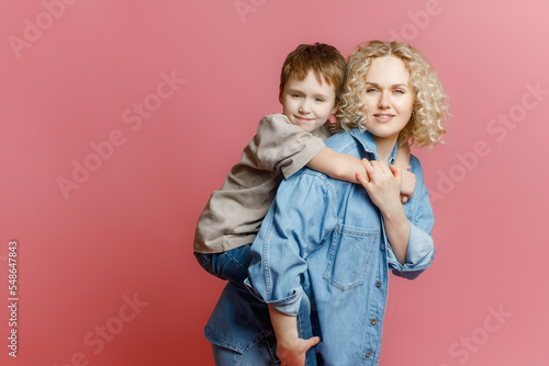 Happy young mother having fun with her child - Son hugs his mom on an isolated pink background - concept of family lifestyle, motherhood, love and tender moments