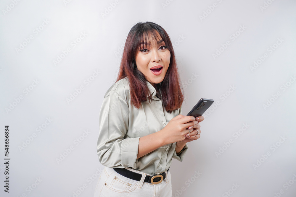 Naklejka premium Shocked Asian woman wearing sage green shirt and holding her phone, isolated by white background
