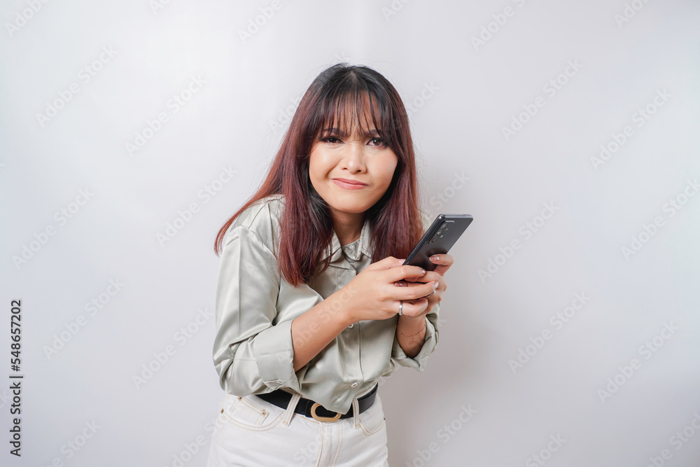 Fototapeta premium A dissatisfied young Asian woman looks disgruntled wearing sage green shirt irritated face expressions holding her phone