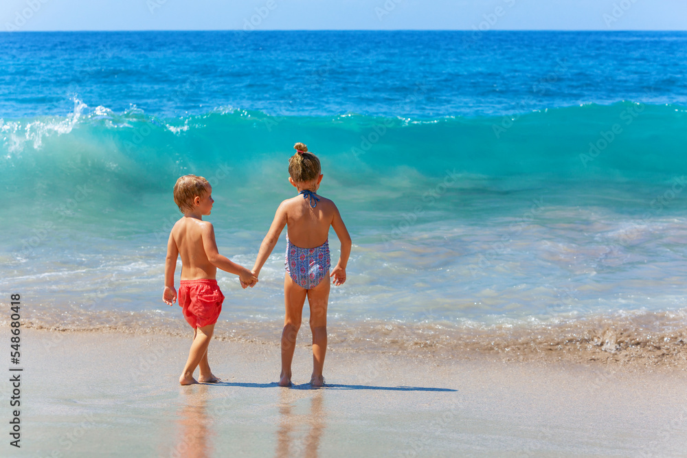 Happy kids have fun in sea surf on white sand beach. Couple of children stand in water pool with hands up. Travel lifestyle, swimming activities in family summer camp. Vacations on tropical island.