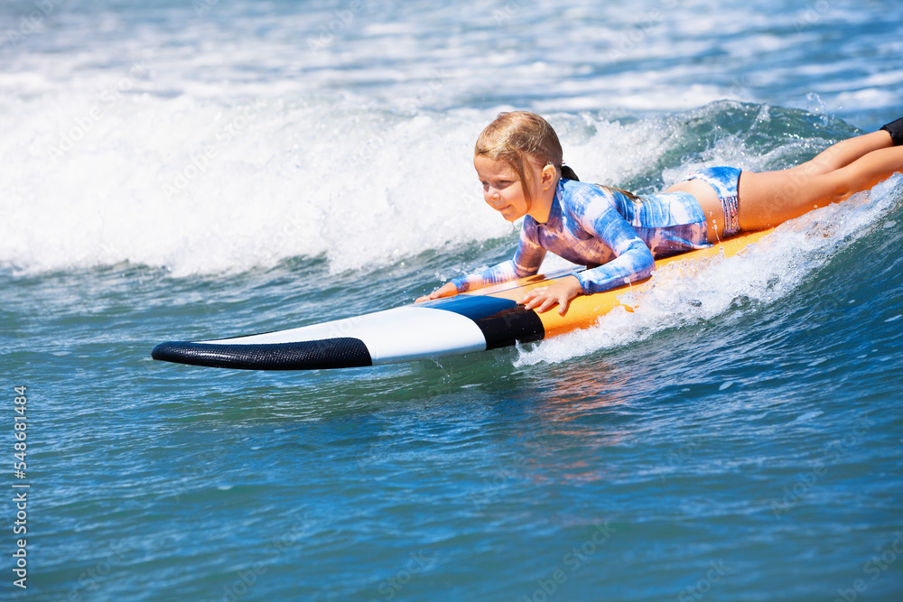 Naklejka premium Happy baby girl - young surfer ride on surfboard with fun on sea waves. Active family lifestyle, kids outdoor water sport lessons and swimming activity in surf camp. Beach summer vacation with child.