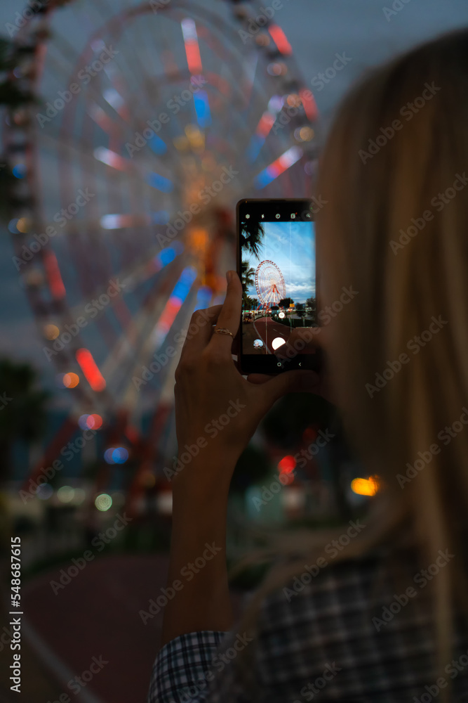 Rear view of a young woman photographing a glowing Ferris wheel on a mobile phone at dusk, sightseeing. Vertical photo