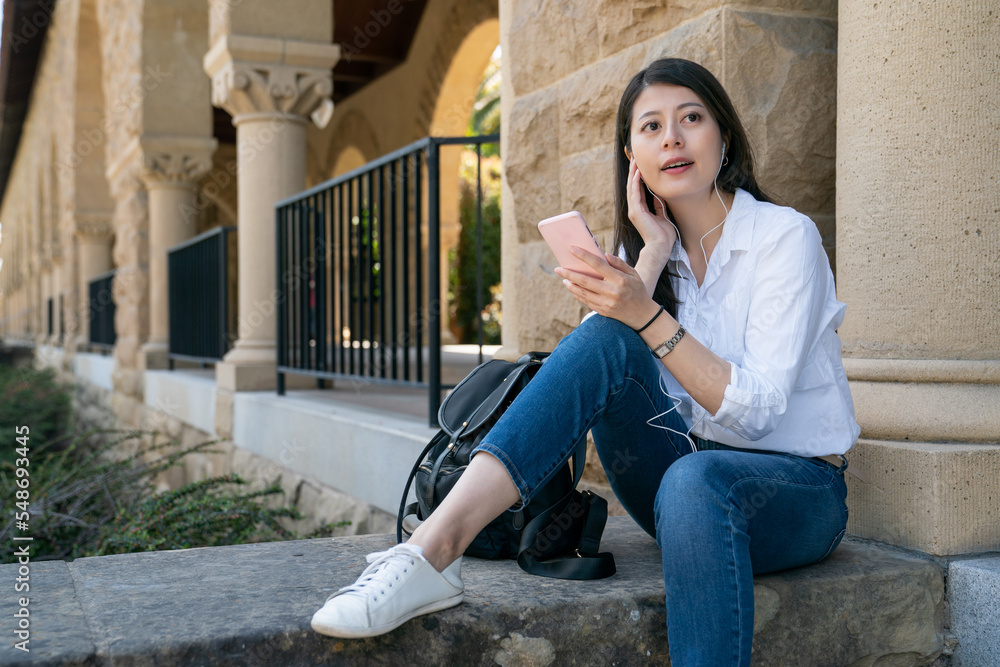 pretty asian Japanese college girl holding smartphone and enjoying music from earphones while sitting relaxing near a hallway at school university in California usa pretty asian Japanese college girl holding smartphone and enjoying music from earphones while sitting relaxing near a hallway at school university in California usa