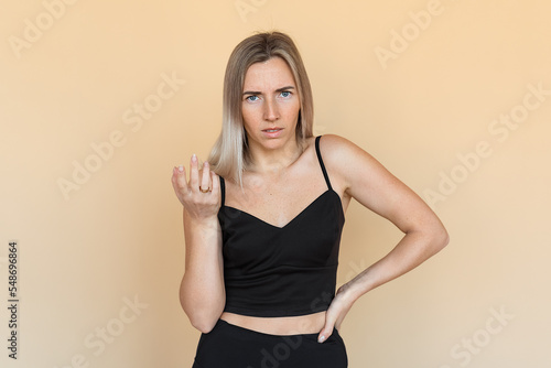 Serious young woman with freckles gestures hand, looking frowning and thoughtful, stands over beige background