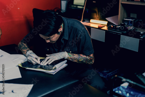 Top view of tattoo artist wearing sterile white gloves while preparing making copy of pattern for tattoo, redrawing it on special paper in a modern studio lowlight.