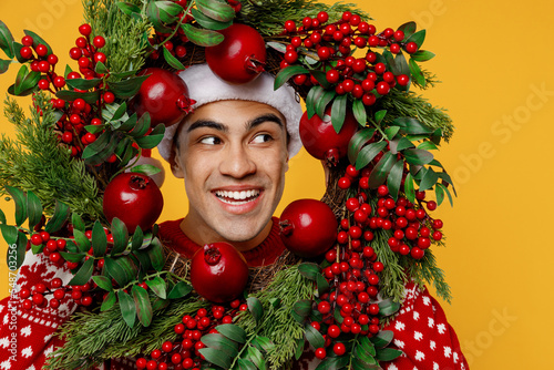 Close up merry smiling young man wear red knitted sweater Santa hat posing hold look aside through Christmas wreath isolated on plain yellow background Happy New Year 2023 celebration holiday concept. © ViDi Studio