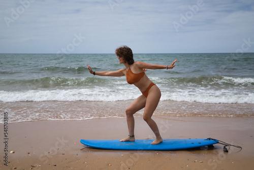 Attractive mature woman with curly hair, sunglasses and bikini, doing funny poses on a blue surfboard on the shore of the beach. Concept sea, sand, sun, beach, vacation, surf, summer.