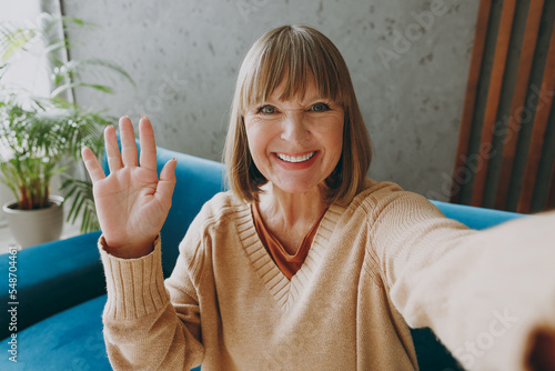 Konstfotografi Close up elderly woman 50s year old wear casual clothes sit on blue sofa do selfie shot pov mobile cell phone waving hand stay at home flat rest relax spend free spare time in living room grey wall