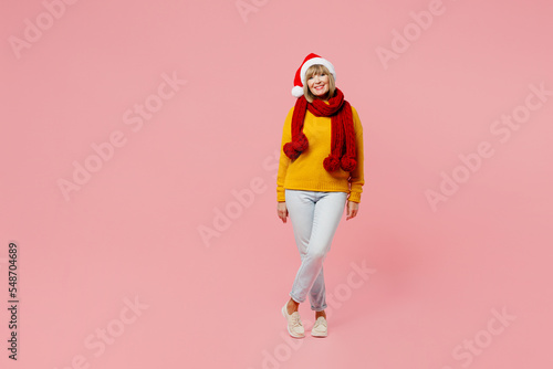 Full body smiling merry cheerful elderly woman 50s years old wear yellow knitted sweater red scarf Santa hat posing look camera isolated on plain pink background Happy New Year Christmas 2023 concept