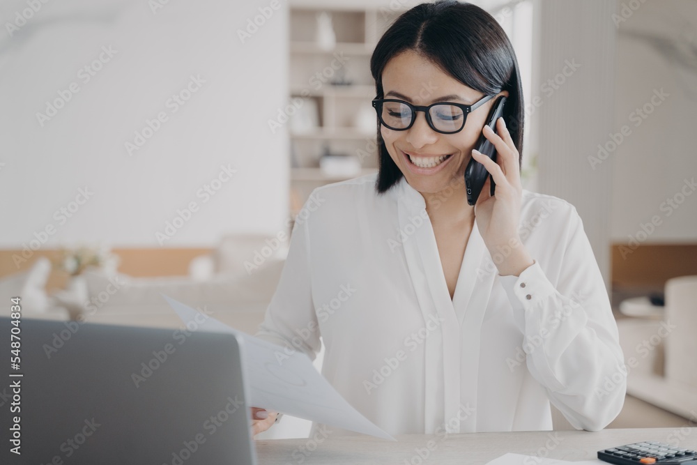 Smiling businesswoman talking on phone, answering call, works with documents, sitting at laptop Smiling businesswoman talking on phone, answering call, works with documents, sitting at laptop
