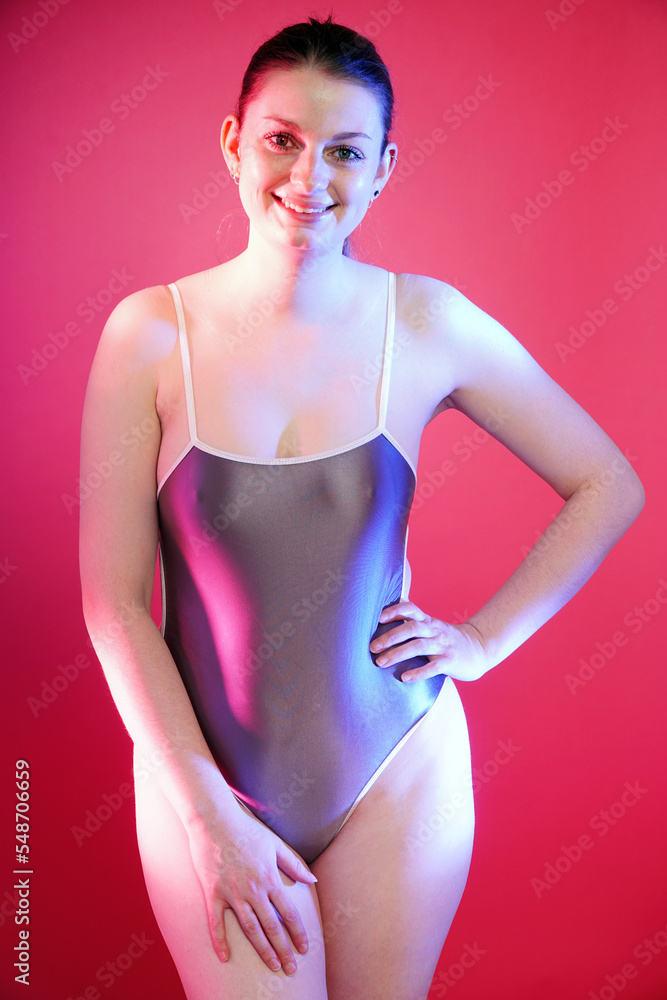 Cheerful young woman posing in silver metallic one piece swimsuit in front of pink background in studio