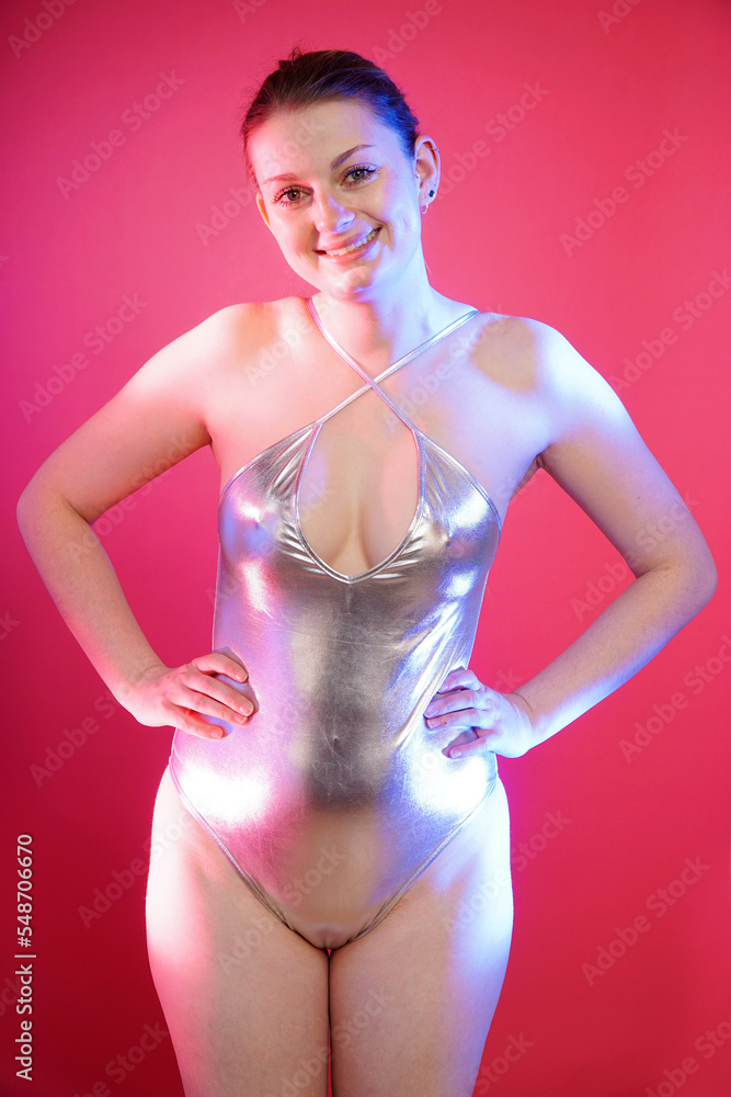 Beautiful cheerful woman posing in silver metallic one piece swimsuit in front of pink background in studio