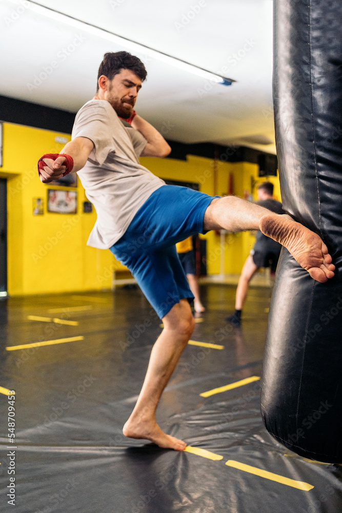 Bearded young man kicking the punching bag in training of kickboxing