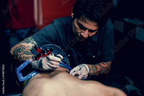 Professional tattooist makes the tattoo on a men waist, focusing on tattoo machines in a modern studio lowlight.