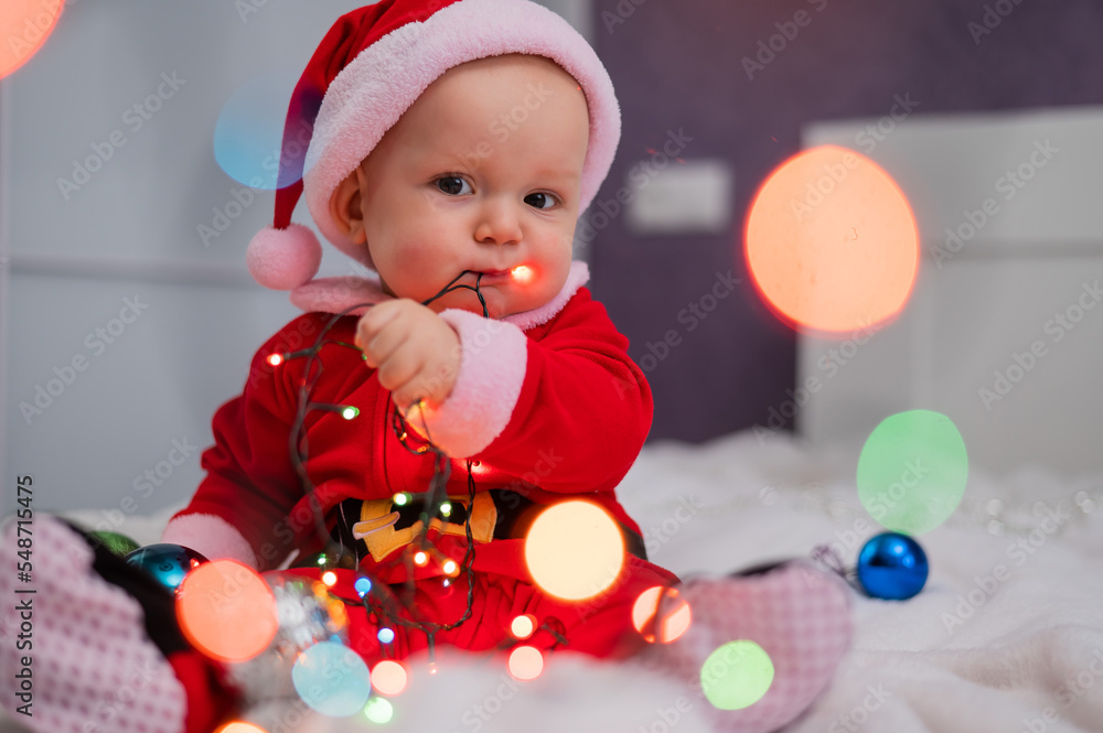 Obraz premium Baby boy wearing in santa claus sits on the bed playing with lights.