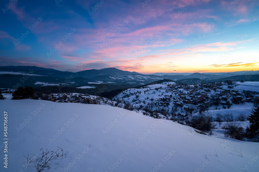 Beautiful sunrise view with snowy mountain slopes and small village among them in the frozen winter morning, the Rhodopi Mountains, Bulgaria .