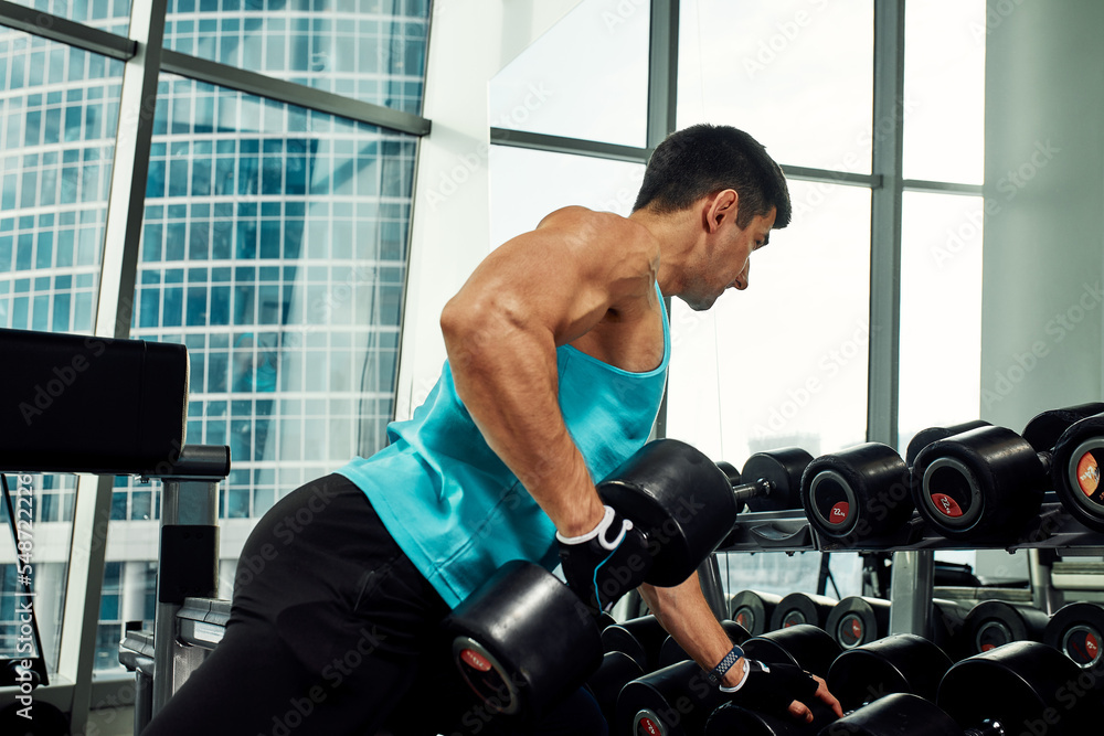 Man doing exercise with dumbbell in the gym. Photo of a sexy muscular man in sportswear and good physique on grey background. Strength and motivation, sport, fitness goal Man doing exercise with dumbbell in the gym. Photo of a sexy muscular man in sportswear and good physique on grey background. Strength and motivation, sport, fitness goal