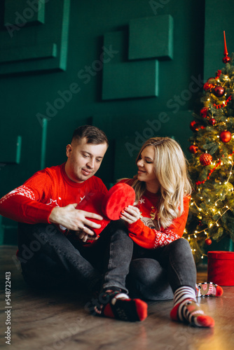 A charming couple in love in traditional Christmas red sweaters spend time near the Christmas tree © anatoliycherkas