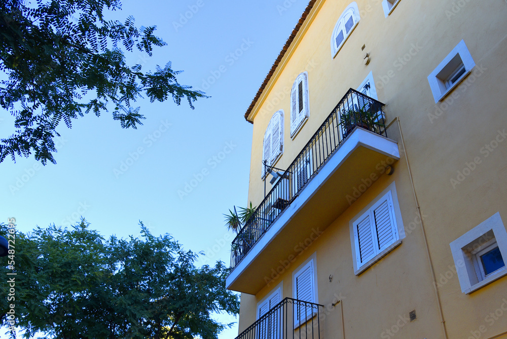 Balcony with windows on a residential building. Yellow building facade against trees with green leaves. Buildings architecture and house facade. Modern minimalistic architecture in Spain. Balcony with windows on a residential building. Yellow building facade against trees with green leaves. Buildings architecture and house facade. Modern minimalistic architecture in Spain.