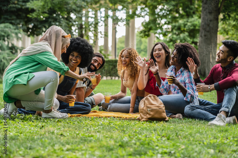 Multiracial group of friends sitting on the grass in a public park drinking orange juice and waving