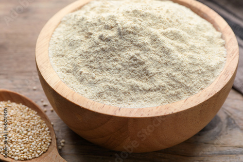 Quinoa flour in bowl and spoon with seeds on wooden table, closeup photo