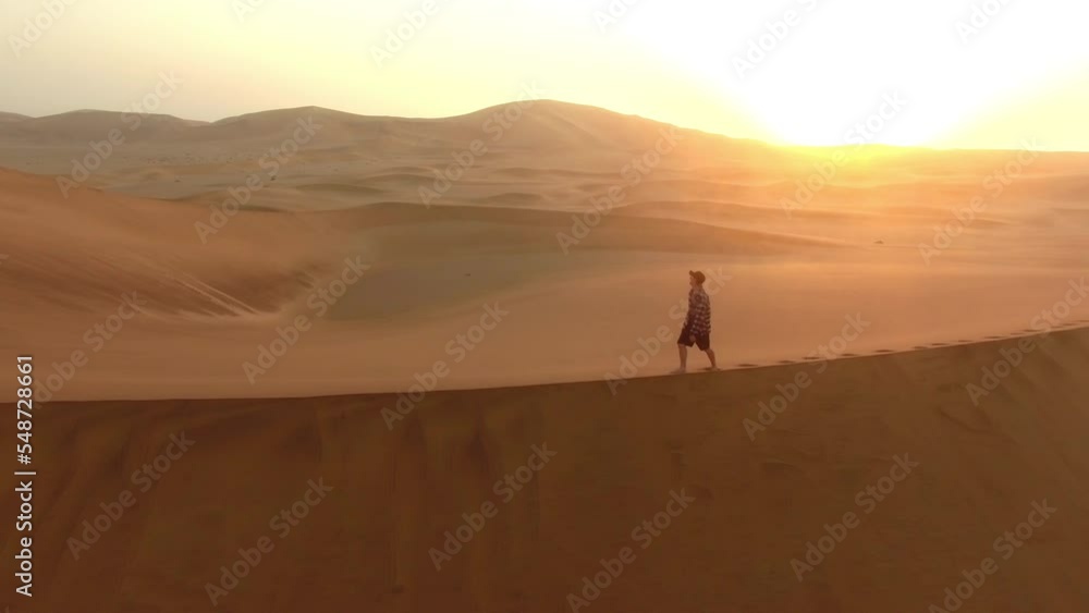 Man, walking and drone in desert or mountains of sand with sunset background. Person, travel and adventure for freedom or nature and gold land, carbon footprint in sand dunes of Namibia in Africa Man, walking and drone in desert or mountains of sand with sunset background. Person, travel and adventure for freedom or nature and gold land, carbon footprint in sand dunes of Namibia in Africa