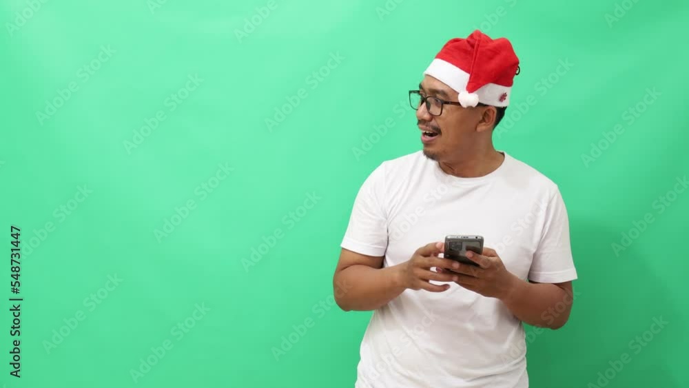 Surprised asian man standing while holding cell phone and pointing sideways. Christmas concept isolated on green background Surprised asian man standing while holding cell phone and pointing sideways. Christmas concept isolated on green background