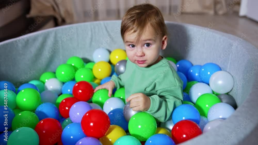 Cute one year old toddler sits in soft basin with plastic balls. Sweet boy looks at the balls calmly. Kid passes a toy to mom and heads to the parent. Cute one year old toddler sits in soft basin with plastic balls. Sweet boy looks at the balls calmly. Kid passes a toy to mom and heads to the parent.