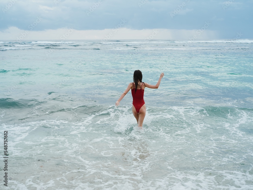 Woman standing with her back to the camera tourist in a red bathing suit sitting on the sand on the beach in the ocean in the waves, travel and beach vacation
