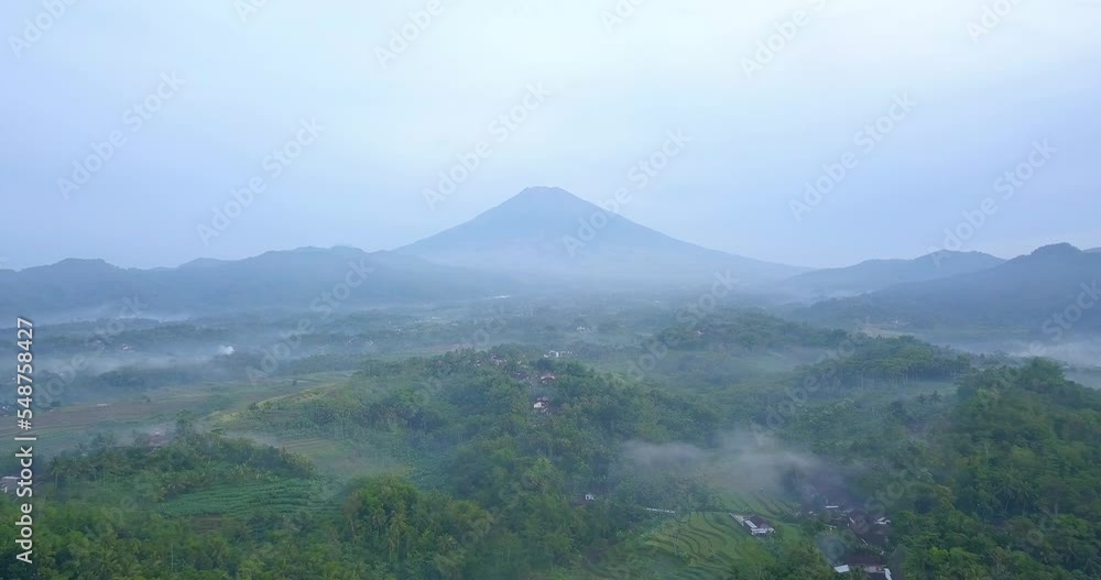 Aerial view of rural landscape in the misty morning with huge mountain on the background. Village and trees shrouded in mist - Tropical countryside of Indonesia Aerial view of rural landscape in the misty morning with huge mountain on the background. Village and trees shrouded in mist - Tropical countryside of Indonesia