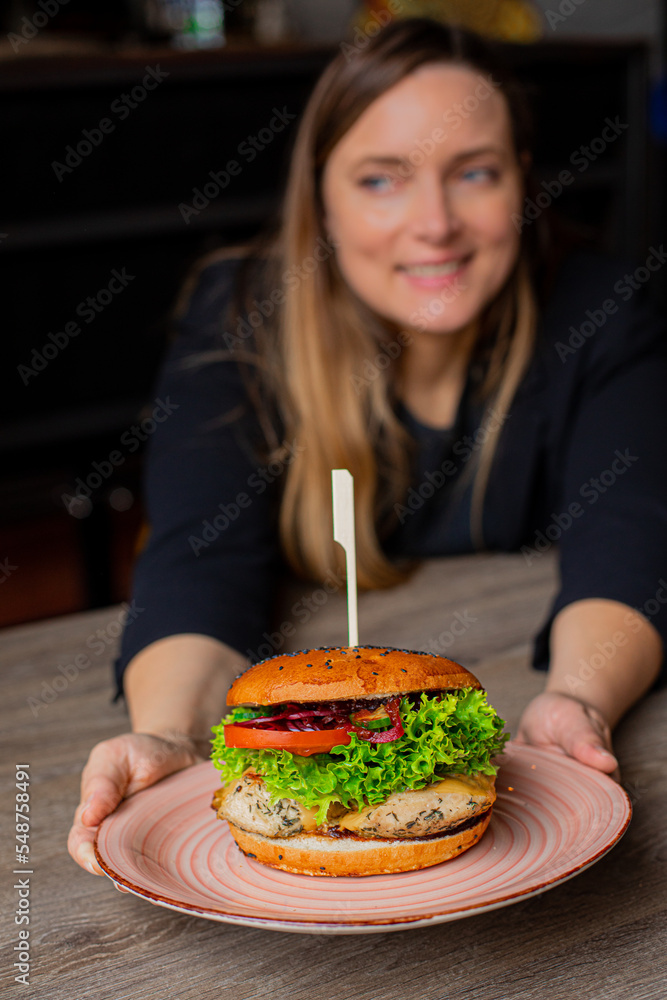 Vertical of blurred woman holding large double hamburger with skewer on pink plate in restaurant or cafe. New taste