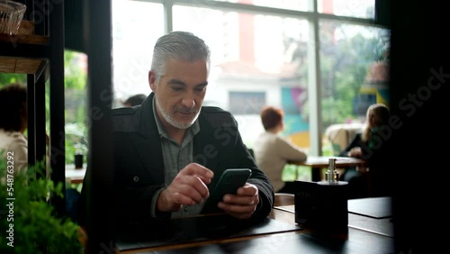 Middle aged entrepreneur working at coffee shop using cellphone modern device. Older person looking at smartphone reading message on phone