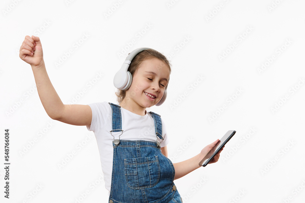 Lovely child, joyful baby girl in blue denim overall, holding smartphone and dancing while listening to the music on stereo headphones, isolated over white background with copy ad space for text Lovely child, joyful baby girl in blue denim overall, holding smartphone and dancing while listening to the music on stereo headphones, isolated over white background with copy ad space for text