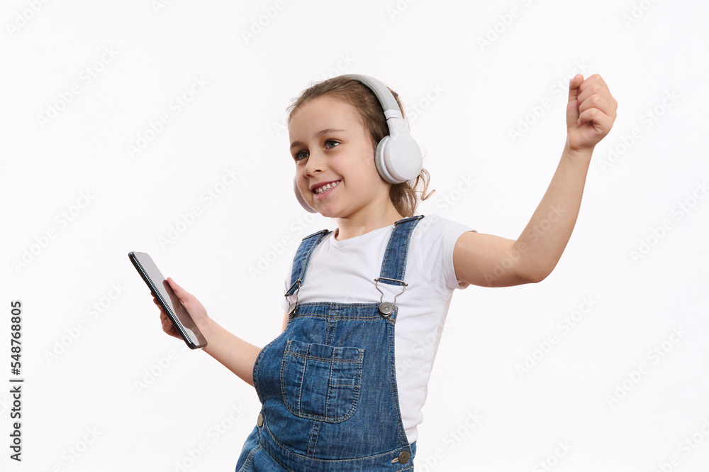 Joyful Caucasian child, a cute little girl wearing blue denim overalls, with smartphone in her hands, dances and listens to the music on her headphones, isolated over white background with copy space Joyful Caucasian child, a cute little girl wearing blue denim overalls, with smartphone in her hands, dances and listens to the music on her headphones, isolated over white background with copy space
