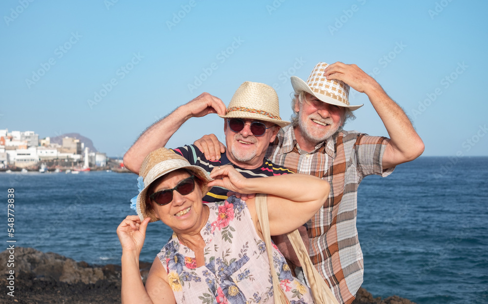 A group of three playful older people holding their hats from flying off enjoying a windy day at sea. Happy retirees on beach vacation. Horizon over water