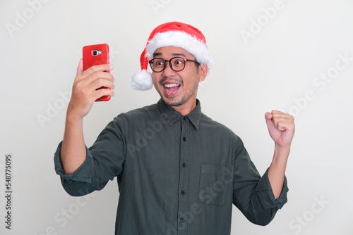 Asian man wearing Christmas hat showing excited when looking to his handphone photo