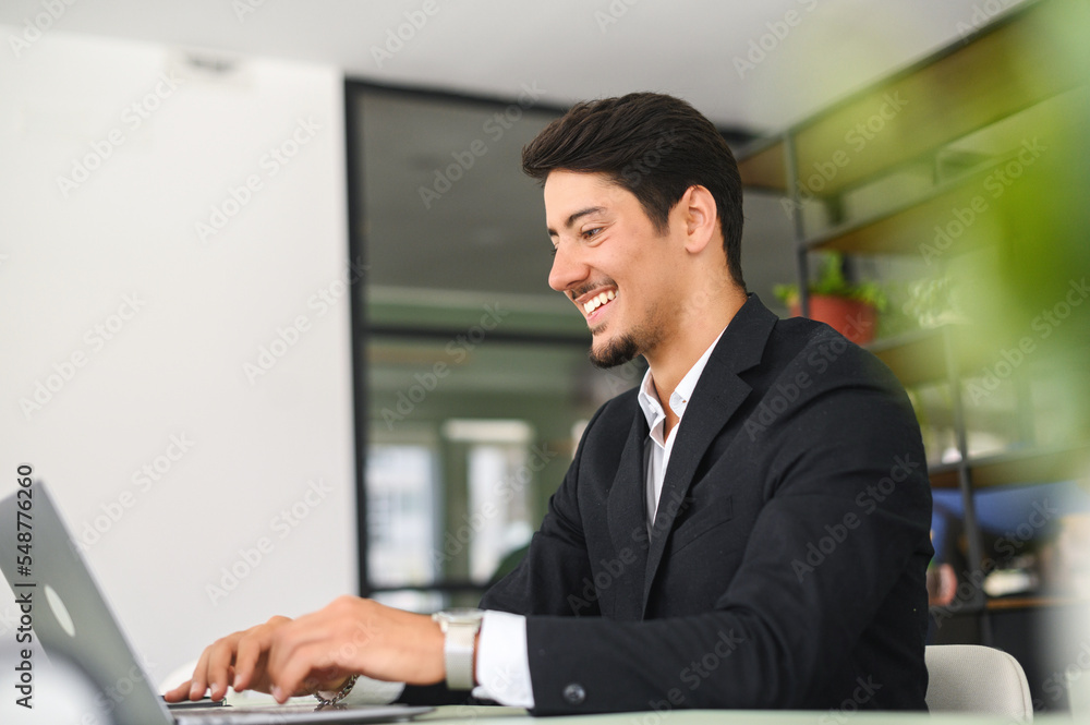 Happy smart young hispanic businessman wearing formal suit using the laptop computer at modern office. Smiling latin male office employee at the workplace is typing Happy smart young hispanic businessman wearing formal suit using the laptop computer at modern office. Smiling latin male office employee at the workplace is typing