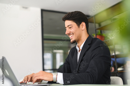 Happy smart young hispanic businessman wearing formal suit using the laptop computer at modern office. Smiling latin male office employee at the workplace is typing © Vadim Pastuh