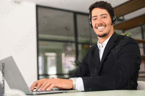 Friendly and smiling hispanic man wearing wireless headset looks at the camera sitting at the desk in front of laptop in modern office, male employee using hands free device for online connection