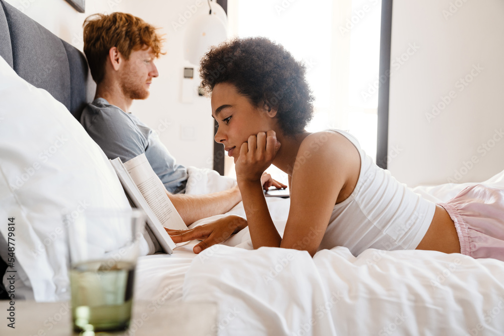 Young beautiful african woman reading lying with her working boyfriend Young beautiful african woman reading lying with her working boyfriend