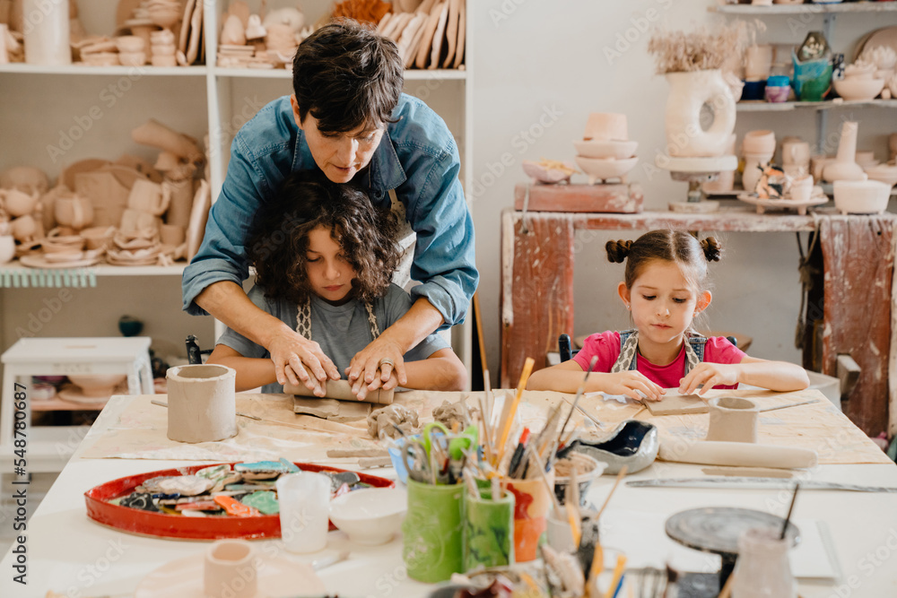 Mature female teacher making pottery crafts with group of kids at workshop