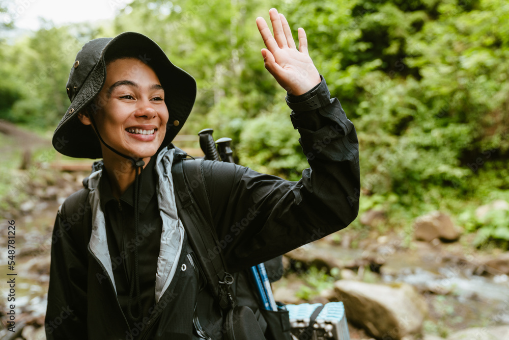 Happy white woman wearing backpack waving while hiking in forest Happy white woman wearing backpack waving while hiking in forest