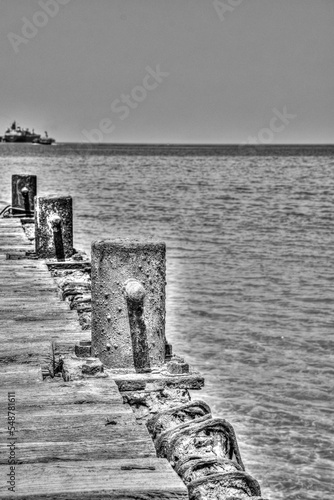 Vertical grayscale of an old wooden dock of a lake, rusty poles on sides and a yacht on the horizon