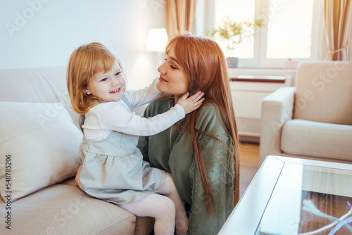 Mother and daughter having fun on sofa. Beautiful young woman and her charming little daughter are hugging and smiling. Happy loving family. Mother and her daughter child girl playing and hugging.