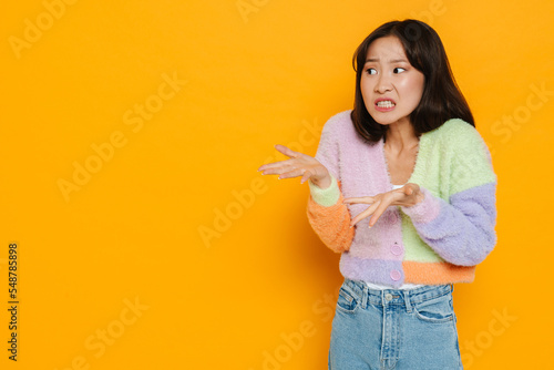 Upset asian girl pointing both hands at copy space isolated over yellow background
